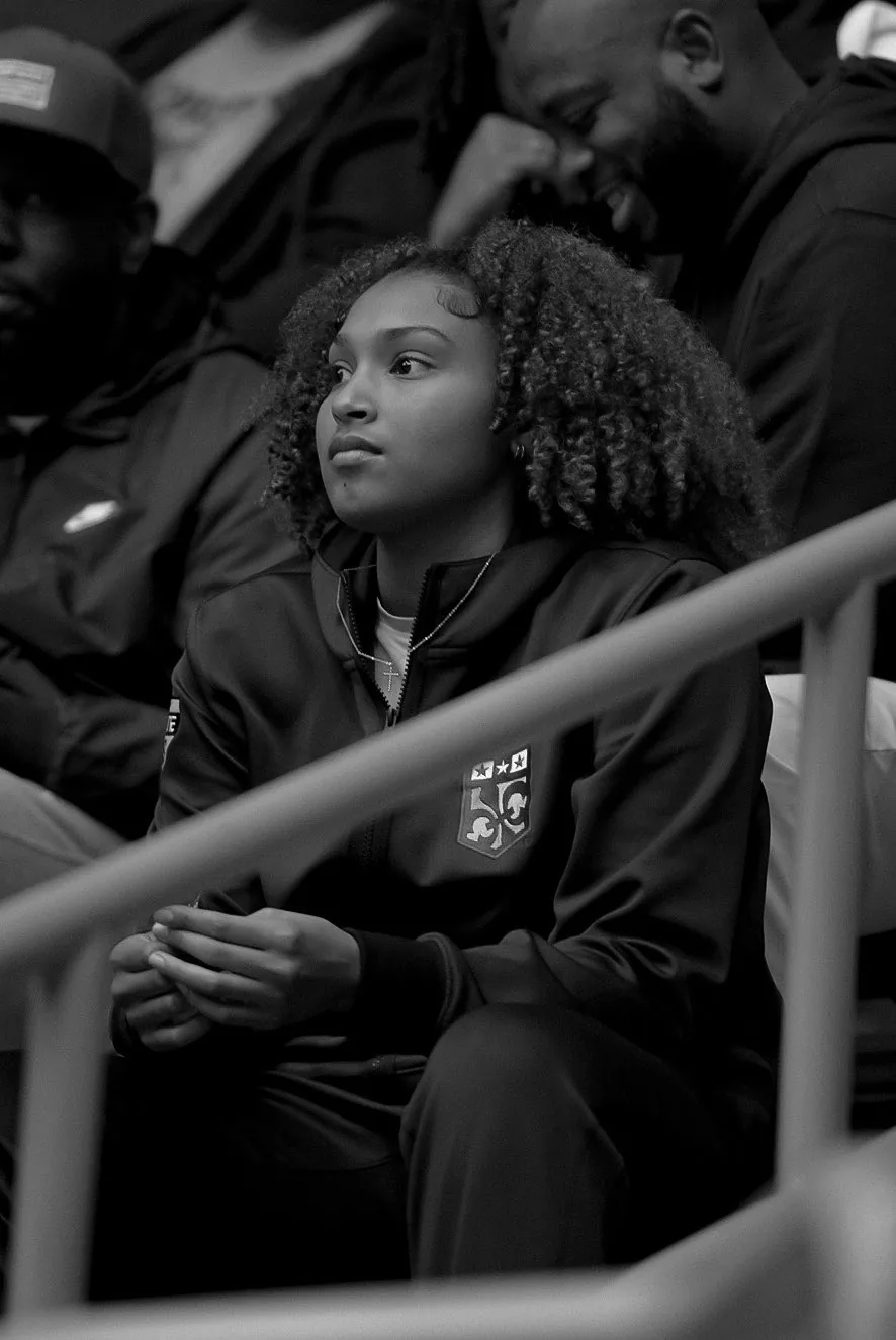 Black and white photo of Amayah Garcia sitting in the stands in a warm-up jacket, watching the floor.
