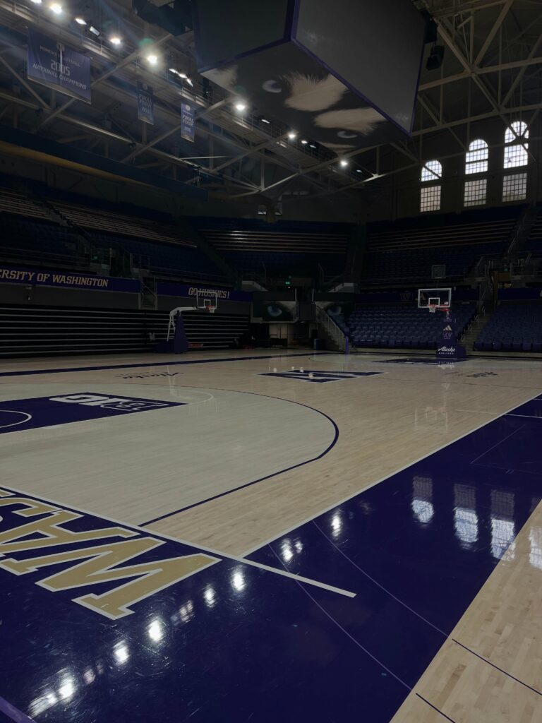Empty basketball court and stands at the University of Washington arena, with championship banners above and Go Huskies painted on the seats