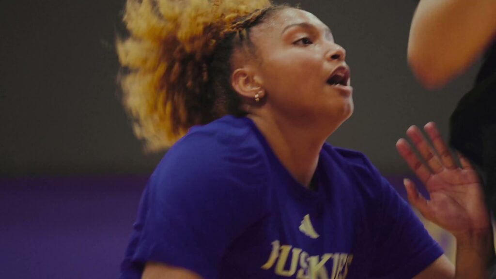 Amayah mid-effort at Washington practice, sweat on her forehead and mouth open catching her breath, in a purple Huskies shirt against the facility wall